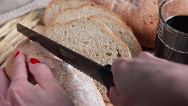 Close-up of a woman slicing fresh rustic bread into pieces with a serrated knife, surrounded by wheat stalks, sliced bread and a dark beverage