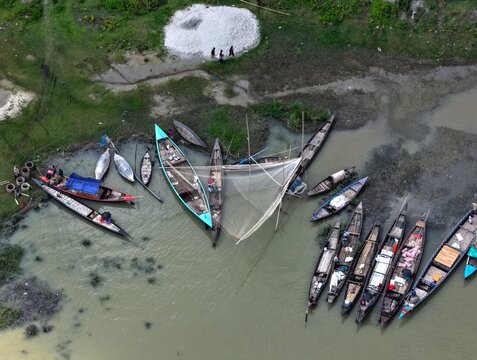 Manikganj, Bangladesh - 27 August 2026: Aerial view of wooden fishing boats moored along the muddy riverbank with fishermen preparing nets and people walking on the grassy shore.