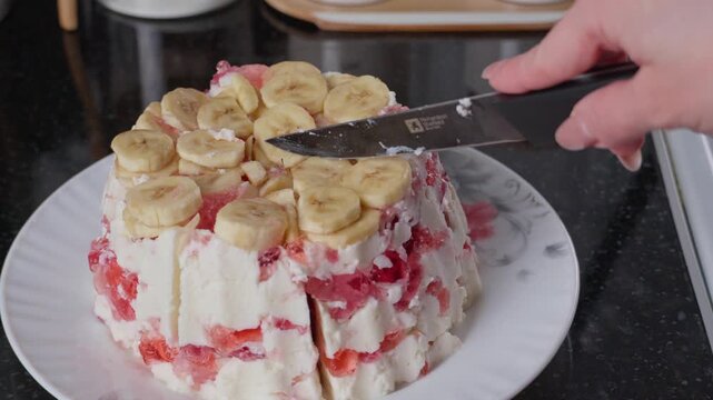 Close-up video of a woman cutting a homemade fruit jelly cake topped with banana slices and layered with creamy filling and red berry jelly