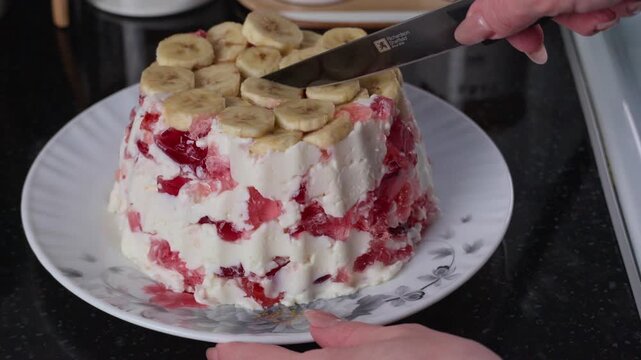 Close-up video of a woman cutting a homemade fruit jelly cake topped with banana slices and layered with creamy filling and red berry jelly