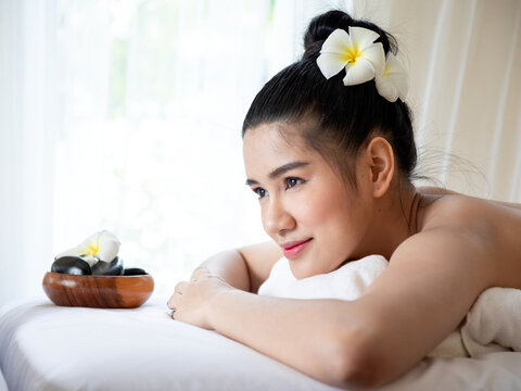Woman relaxing at spa with flower in hair, towel, massage stones and peaceful expression, representing wellness, beauty, massage therapy, comfort and self care