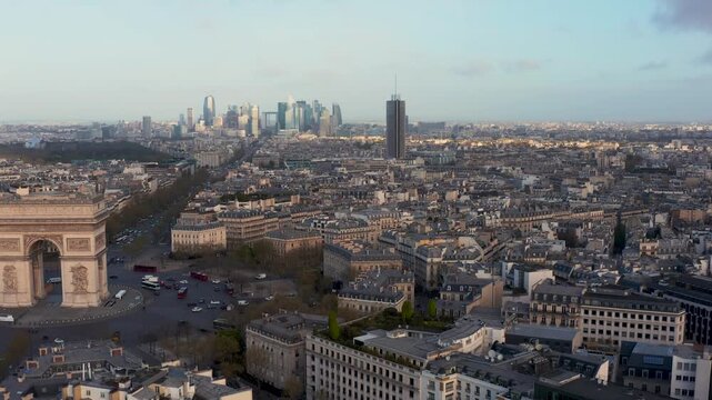 Aerial view of the iconic Arc de Triomphe and the dense urban landscape of Paris with the distant La Defense business district under soft golden hour light, Paris, Ile-de-France, France.