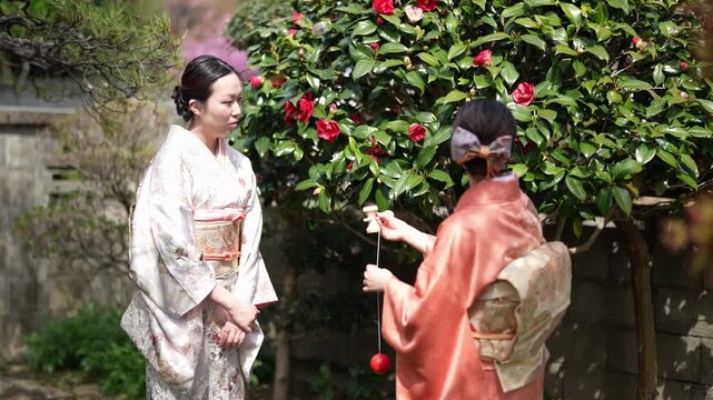 A woman in an orange kimono holds a wooden kendama with its red ball hanging from a string beside another woman. Japan, April 19, 2025, spring.