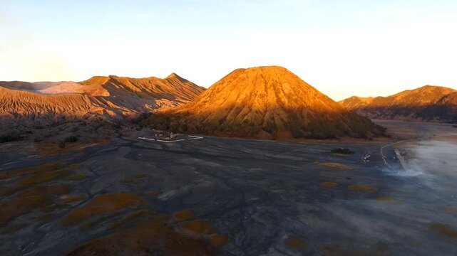 Aerial view of the volcanic landscape of Gunung Batok at sunrise with golden light on the slopes and a sandy caldera floor in Kecamatan Tosari, Jawa Timur, Indonesia.