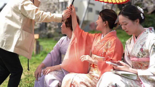 A seated woman in an orange kimono receives an open paper parasol beside other women in kimonos. Japan, April 19, 2025, spring.