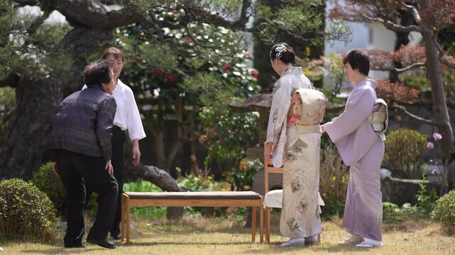 Women in kimonos and casual clothing gather beside a low bench under a pruned tree in a traditional garden. Japan, April 19, 2025, spring.