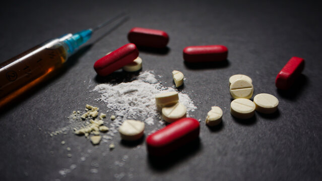 Prescription drugs in pills and powder form with red and white colors on black background. There is a syringe full with brown liquid behind it. Misused concept of drugs. 