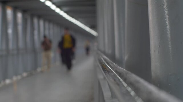 Blurred Pedestrians Walking Through Elevated Skywalk Corridor with Sharp Metal Railing Foreground