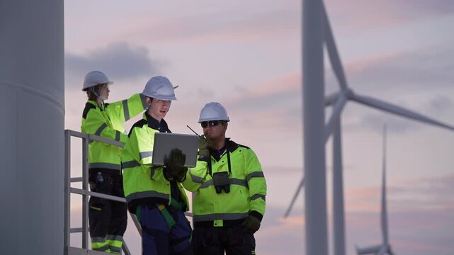 Engineers wearing safety helmets and jackets inspecting a wind turbine at wind farm at sunset. Concept renewable energy, sustainable technology wind power maintenance and environmental engineering.