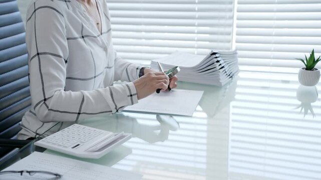 Professional is meticulously examining financial documents with a magnifying glass while writing notes. Surrounded by stacks of paperwork and a calculator on a glass desk