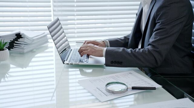 Business professional hands typing on a laptop keyboard. Working on financial documents. With a magnifying glass nearby. Representing analysis. Auditing