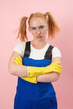 Young woman with red hair and a displeased expression, wearing blue overalls, yellow rubber gloves, and protective eyewear, crossing her arms against a pink background