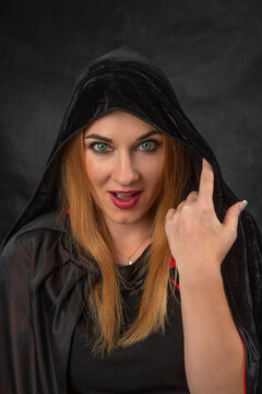 Woman with striking green eyes and red lips looking directly at the viewer, wearing a mysterious black hooded cloak, evoking themes of magic, Halloween, and mystical allure