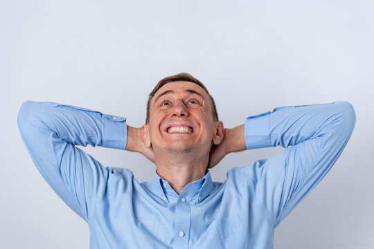 Cheerful adult man in a blue shirt is relaxing with his hands behind his head, smiling broadly and looking upwards with a hopeful expression against a light background