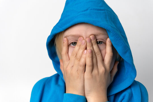 Young child wearing a blue hoodie covering face with both hands, looking through spread fingers with a shy or fearful expression against a plain white background