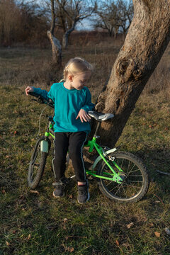 Young blonde girl standing next to her green bicycle, holding the handlebars while resting against a tree in a sunny outdoor setting, learning new skills