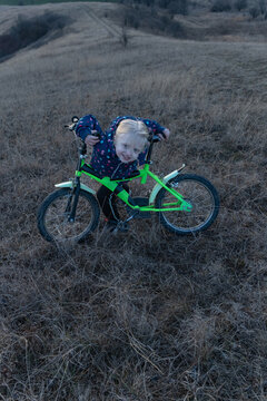 Young child smiles at camera while holding a green bicycle on a dry grassy hill, dressed for cool weather during a cloudy autumn day, radiating joy and countryside playfulness