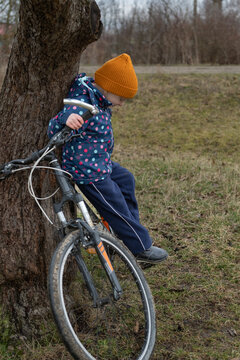 Young child wearing a yellow beanie and blue jacket leaning on a tree trunk, holding a bicycle handlebar, pausing during an outdoor activity on a grassy field