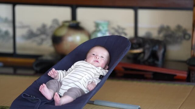 A baby in striped clothing reclines with bare feet in a navy bouncer on a tatami floor. Japan, April 19, 2025, spring.