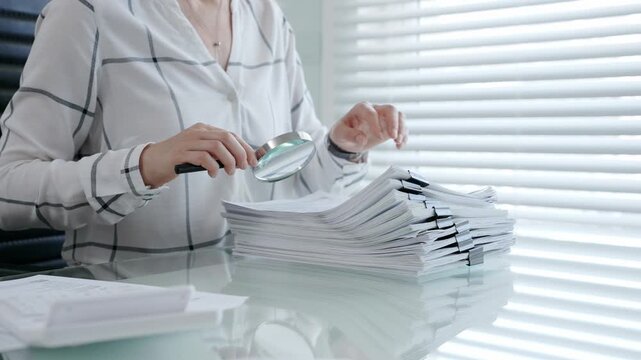 Person examining documents with a magnifying glass, performing detailed financial audit, administrative investigation, legal research, and thorough paperwork inspection in an office