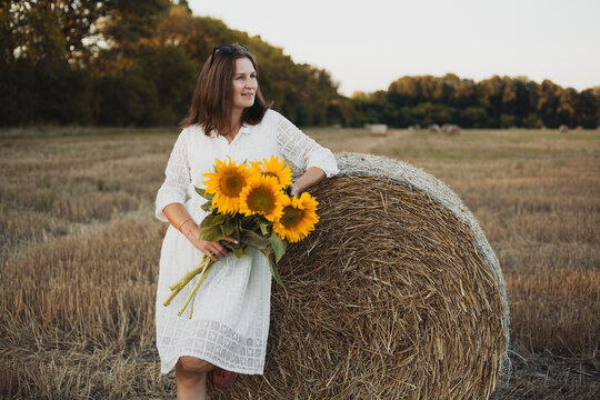 woman in a white dress holding a bouquet of wild flowers with sunflowers in a mowed field with straw bales