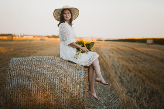 woman in a white dress holding a bouquet of wild flowers with sunflowers in a mowed field with straw bales
