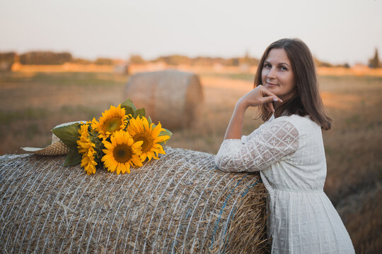 woman in a white dress holding a bouquet of wild flowers with sunflowers in a mowed field with straw bales