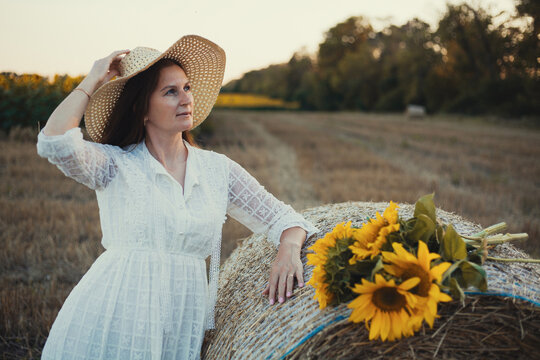 woman in a white dress holding a bouquet of wild flowers with sunflowers in a mowed field with straw bales