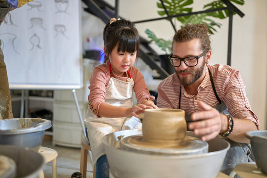 happy girl shaping clay pot on wheel next to the teacher at pottery workshop.  Creative, hobby ,workshop  concept