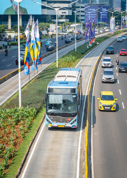 Jakarta, Indonesia - September 6, 2026: Traffic on Jalan Sudirman featuring a TransJakarta bus, private vehicles, roadside flags, landscaped areas, and high-rise buildings in the background.