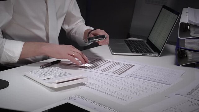 Person working late in an office, meticulously scrutinizing paperwork with a magnifying glass and pen, surrounded by a laptop, calculator, and binders filled with documents