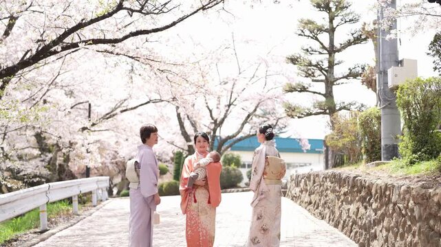 Three women in kimonos look back on a paved street beneath cherry blossoms, with the woman in orange holding a baby. Japan, April 19, 2025, spring.