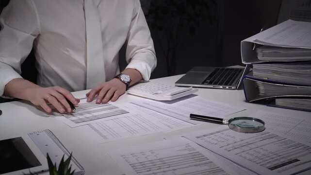 Person working late in an office, meticulously scrutinizing paperwork with a magnifying glass and pen, surrounded by a laptop, calculator, and binders filled with documents