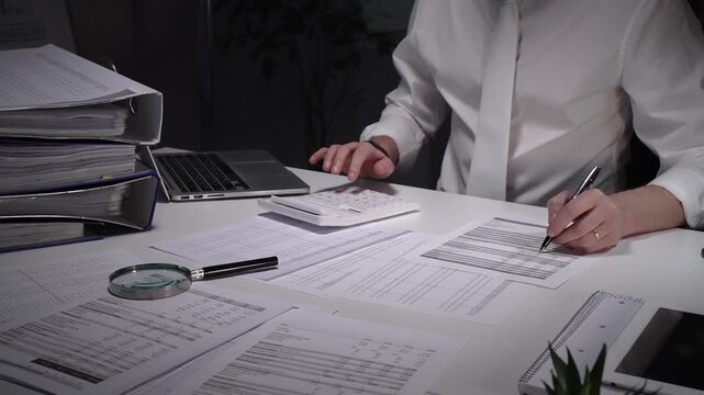 Person working late in an office, meticulously scrutinizing paperwork with a magnifying glass and pen, surrounded by a laptop, calculator, and binders filled with documents