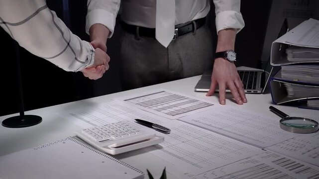 Business professionals shake hands over a cluttered desk of contracts, calculator and magnifying glass, sealing a trusted partnership after detailed financial review and late night work
