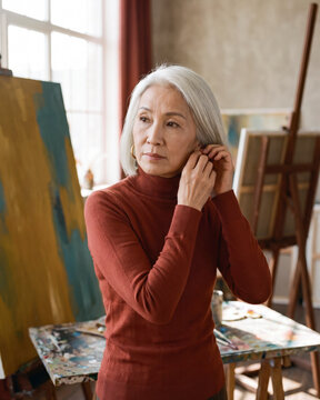 Confident mature artist adjusting earring beside studio easel with warm natural light