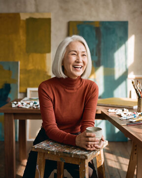 Engaged mature artist laughing on stool in painting studio with warm natural light