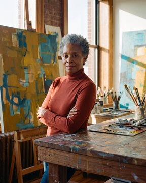Focused mature artist standing beside easel in sunlit painting studio with warm natural light
