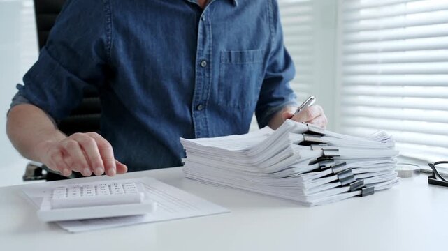 Individual working diligently on financial documents, calculating expenses and reviewing reports at an office desk, managing a large stack of paperwork and invoices