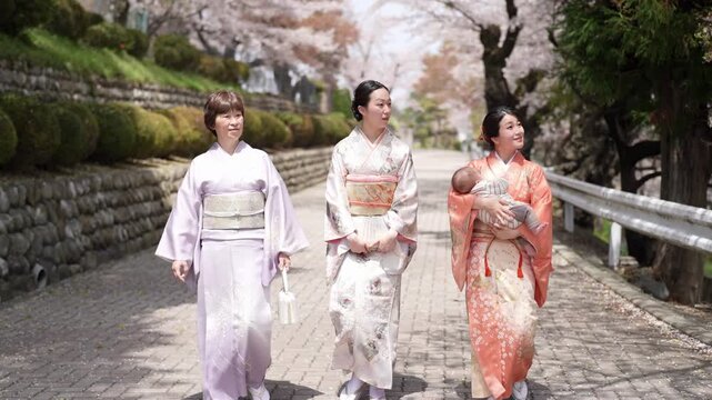 Three women in lavender, patterned and orange kimonos walk along a paved street, with a baby held by the woman in orange. Japan, April 19, 2025, spring.