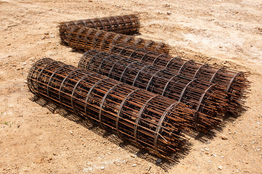 Rolled reinforcement mesh coils rest closely together on sandy ground, welded wire netting awaits use in upcoming concrete slab construction, closeup view highlights sturdy metal grid pattern