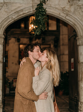 Romantic young couple kissing passionately under a historic stone arch decorated with festive greenery