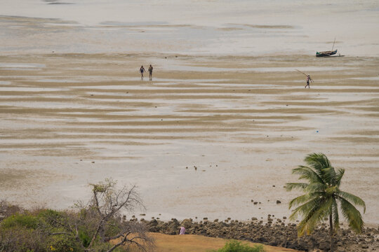 Antsiranana, Diana, Madagascar - 15 June 2026: View of people walking across the vast mudflats during low tide with a small boat and a palm tree along the rocky coast.