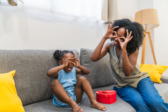 African american mother and daughter having fun on a couch, making funny faces and celebrating a special occasion with a gift box