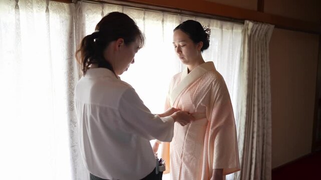 An assistant ties a waist sash around a woman wearing a peach kimono underlayer by the window. Japan, April 19, 2025, spring.
