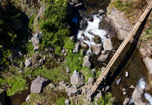 Aerial Scenery of Traditional Wooden Bridge and Water Pipeline Crossing Above a Clear Rocky River in Indonesia.