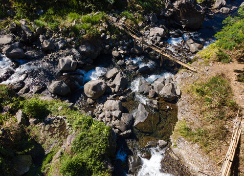 Aerial Scenery of Traditional Wooden Bridge and Water Pipeline Crossing Above a Clear Rocky River in Indonesia.
