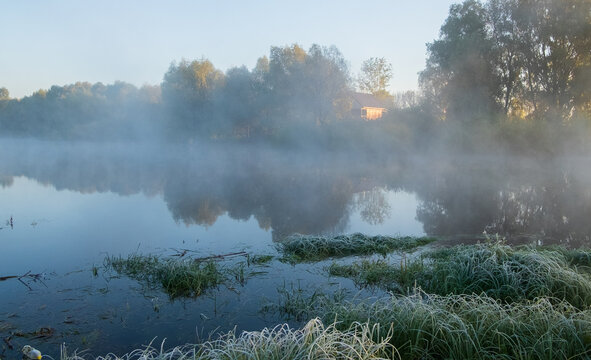 A calm river reflects the morning sun and thick fog rolling over the grassy banks and distant autumn trees.