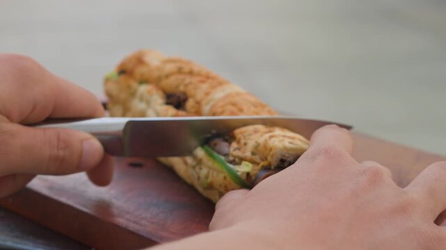 Close-up side angle of person hands slicing a fresh long submarine sandwich in half with a kitchen knife on a wooden cutting board to reveal the inside fillings, horizontal