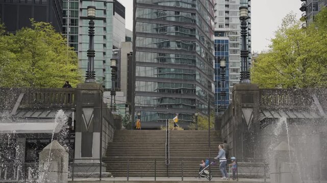 People walking down steps in urban park near water fountain in front of tall skyscrapers.